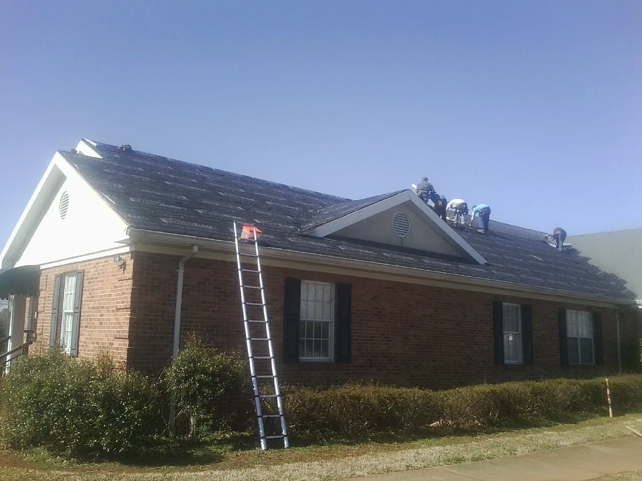 Crew tearing shingles off a brick ranch roof with a ladder set — Emergency Roof Repair work by our crew in Calabash