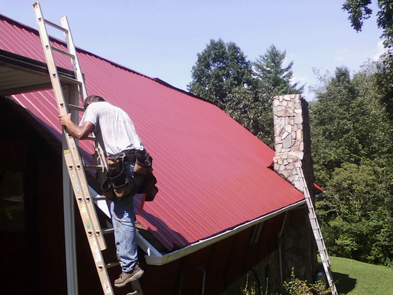 Roofer on a ladder beside a red metal roof and stone chimney — Soffit & Fascia Repair by Roofing Brothers in Calabash